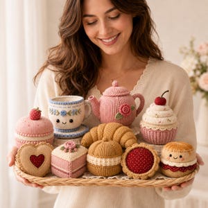 Könnte beinhalten: Eine Sammlung von gestrickten Spiellebensmitteln auf einem Weidentablett, darunter eine rosa Teekanne mit Rose, eine blau-weiße Teetasse, ein Croissant, ein Cupcake mit Kirsche, ein herzförmiger Keks, ein quadratischer Kuchen und kleine Gebäckstücke.