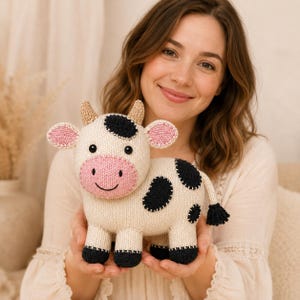 Puede incluir: Un juguete de vaca tejido en color crema con manchas negras en el cuerpo y la cabeza, un hocico rosa y pequeños cuernos de color canela. La vaca tiene ojos negros bordados, una boca cosida, pezuñas negras y una cola con borla negra.