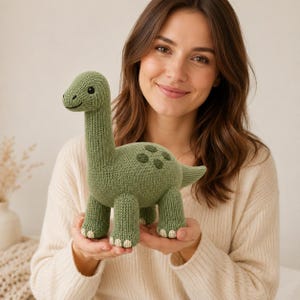 May include: A sage green knitted dinosaur toy featuring a long neck, a rounded body with darker green circular spots on the back, and four sturdy legs with cream-coloured stitched details on the feet. The toy has small black bead eyes.