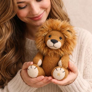 Puede incluir: Un peluche de león tejido, con cuerpo marrón, melena esponjosa y hocico blanco. El león tiene ojos negros y nariz negra. Las patas son color canela con la parte inferior blanca y un detalle de estrella dorada. El juguete se sostiene en las manos.
