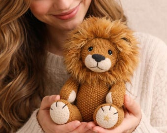 Puede incluir: Un peluche de león tejido, con cuerpo marrón, melena esponjosa y hocico blanco. El león tiene ojos negros y nariz negra. Las patas son color canela con la parte inferior blanca y un detalle de estrella dorada. El juguete se sostiene en las manos.