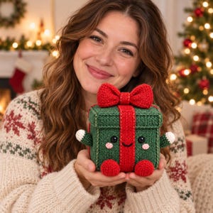 May include: A square, dark green crocheted gift box with a bright red ribbon and a large red bow on top. The box features two small green arms with white tips, two red feet, and a smiling face with black eyes and pink cheeks embroidered on the front.