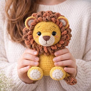 Może przedstawiać: Ręcznie robiona szydełkowa pluszowa zabawka w kształcie lwa. Lew ma żółte ciało, brązową i pomarańczową grzywę oraz biały pysk. Oczy lwa są czarne i ma małe różowe policzki. Lew jest trzymany przez osobę ubraną w biały sweter.