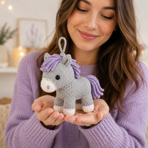 May include: A small, hand-knitted grey horse toy featuring a light purple mane and tail, a white muzzle with a small purple dot, and white hooves. The plush animal has a small loop attached to the top of its head for hanging purposes.