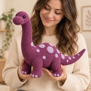 Puede incluir: Un juguete de dinosaurio tejido a ganchillo en hilo morado oscuro con un cuello largo, un cuerpo redondeado y una cola. El dinosaurio tiene manchas circulares de color morado claro en la espalda y los costados, detalles de dedos morados y dos ojos de plástico negro.