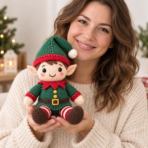 Può includere: Una bambola elfo lavorata a maglia con un cappello verde con pompon bianco, una tunica verde con cintura marrone e fibbia gialla, e gambe a righe rosse e bianche con scarpe marroni. La bambola ha un viso beige e guance rosa.