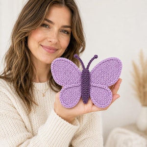 May include: A handcrafted knitted butterfly ornament featuring light purple wings with a darker purple central body and two small antennae. The butterfly is constructed with a visible knit texture and is held in a flat position to display its symmetrical shape.
