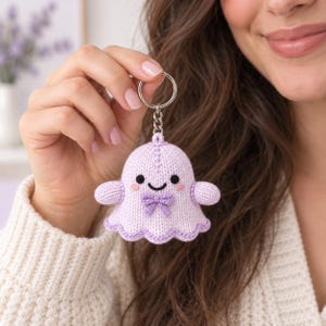 May include: A light purple crocheted ghost keyring featuring a small bow at the neck, black embroidered eyes, a smiling mouth, and pink blush accents. The ghost is attached to a silver-toned metal split ring by a short chain link connector.