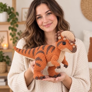 Puede incluir: Un juguete de peluche de dinosaurio paquicefalosaurio tejido a ganchillo con un cuerpo naranja texturizado con rayas marrón oscuro, una cabeza abovedada de color beige claro con pequeñas protuberancias marrones, garras marrones y ojos negros brillantes.