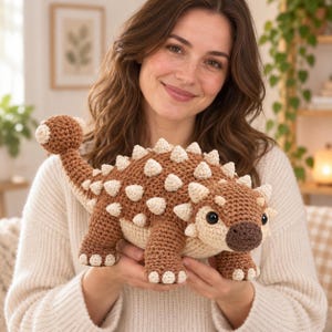 Peut inclure: Un jouet dinosaure ankylosaure au crochet de couleur marron et crème, doté d'un corps texturé avec de petites pointes sur le dos, d'une queue en massue, d'un museau marron foncé et de deux yeux en plastique noir. Fabriqué avec des mailles visibles.