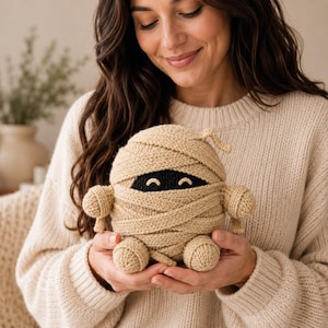 May include: A round, beige, crocheted mummy plush toy featuring a black eye band with two embroidered smiling eyes. The figure has small, rounded arms and legs, and is wrapped in textured, yarn-based bandages that cover its entire body.