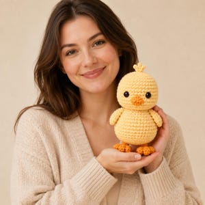 May include: A small, round, yellow crocheted chick toy with a textured yarn surface, featuring two black plastic eyes, a small orange beak, and two orange feet. The chick has a tiny tuft of yellow yarn on the top of its head and a round body.