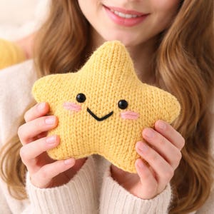 May include: A yellow knitted star-shaped plush toy with a smiling face. The toy has black eyes, a black stitched mouth, and pink blush on its cheeks. The star is held in a person's hands, and the background is blurred.