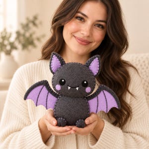May include: A dark gray felt bat plush toy featuring light purple inner ears and wing accents. The bat has black circular eyes, two small white fangs, and round pink cheeks. The edges of the felt are joined with visible light gray stitching.