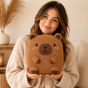 Peut inclure: Une peluche tricotée marron en forme de capybara avec un museau marron foncé, de petites oreilles arrondies, des yeux noirs et des détails de joues roses brodés. Le jouet présente une surface en tricot texturé avec des accents marron foncé.