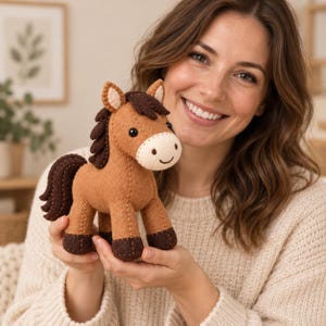 Puede incluir: Un peluche de caballo de fieltro marrón con crin, cola y cascos de color marrón oscuro. El caballo presenta un hocico de color crema con una sonrisa bordada, dos pequeños ojos de cuentas negras y dos orejas puntiagudas con detalles crema.