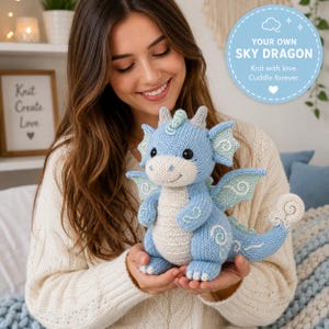 May include: A light blue and cream knitted dragon plush toy with embroidered white swirl details on its wings and tail, featuring two small horns, black eyes, and a stitched smile. The text reads: YOUR OWN SKY DRAGON Knit with love. Cuddle forever.