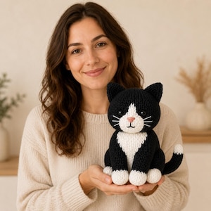 Könnte beinhalten: Ein gehäkeltes Katzen-Plüschtier in Schwarz und Weiß mit schwarzem Körper, weißer Brust, weißen Pfoten und einer rosa Nase. Die Katze hat spitze Ohren, schwarze Augen und weiße gestickte Schnurrhaare und sitzt mit aufgerolltem Schwanz.