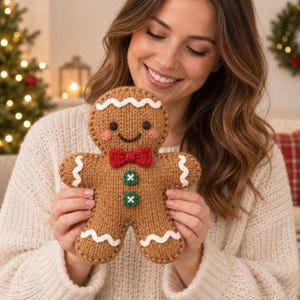 May include: A brown knitted gingerbread man plush toy featuring a red bow tie, two green buttons with white cross-stitching, pink cheeks, and white wavy icing-style trim on the head, arms, and legs, with dark brown embroidered eyes and mouth.