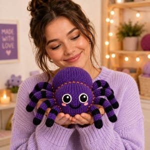 Puede incluir: Un juguete de peluche de araña tejido con un cuerpo de color morado vibrante, patas con rayas negras, ojos grandes en blanco y negro y una pequeña sonrisa bordada. La araña tiene costuras decorativas naranjas en la parte superior de su cabeza redonda.