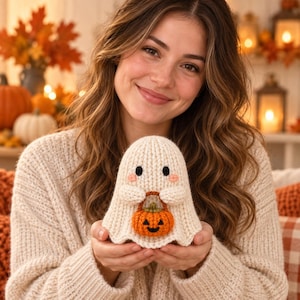 May include: A cream-colored crocheted ghost figurine featuring two black circular eyes, small pink stitched cheeks, and a small orange crocheted pumpkin bucket with a carved jack-o'-lantern face held in its two small rounded arms.