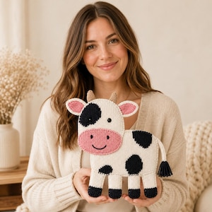 Puede incluir: Un juguete de vaca tejido plano con cuerpo blanco y manchas negras, hocico rosa y pequeños cuernos marrones. La vaca tiene ojos negros, una sonrisa bordada y una borla negra en la cola. Las cuatro patas tienen puntas negras y orejas rosas.
