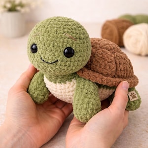 May include: A crocheted turtle toy in green, brown, and cream. The turtle has a smiling face, black eyes, and a textured shell. The toy is held in two hands.