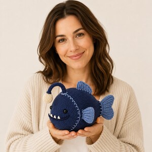 Puede incluir: Un peluche de rape tejido en color azul oscuro con un señuelo beige claro, ojos negros pequeños y dientes triangulares blancos. El pez tiene aletas y cola de color azul claro, con costuras blancas visibles a lo largo del cuerpo.
