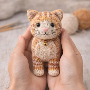 May include: A handmade, knitted cat figurine in shades of orange, tan, and cream. The cat has a pink nose, black whiskers, and a small gold bell collar. The cat is held in two hands, with yarn balls in the background.