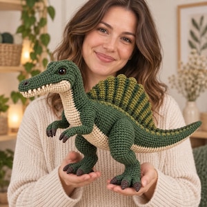 May include: A crocheted Spinosaurus dinosaur toy featuring a dark green body with a lighter beige underbelly and snout. The plush figure has a prominent dorsal sail, dark brown claws, and dark eyes, crafted with visible yarn stitches throughout.