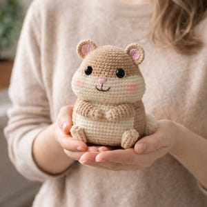 Puede incluir: Un juguete de hámster tejido a ganchillo en tonos beige y crema, con detalles rosas en las orejas y las mejillas. El peluche hecho a mano tiene ojos negros y una pequeña nariz rosa, y se sostiene en manos abiertas. Es un artículo lindo y tierno.