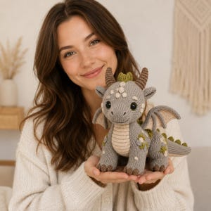 Puede incluir: Un peluche de dragón tejido en color gris con vientre crema, cuernos marrones y pequeños detalles verdes en la espalda y las alas. La criatura tiene grandes ojos negros, una superficie texturizada y un patrón detallado de círculos en la cabeza.