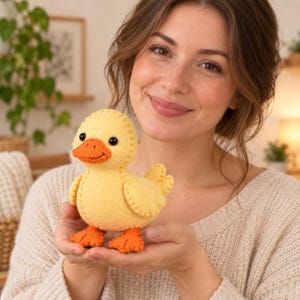May include: A small, handmade felt duck plush toy with a pale yellow body, bright orange beak, and orange webbed feet. The toy features visible blanket stitching along the seams, small black bead eyes, and a stitched smile on the beak.