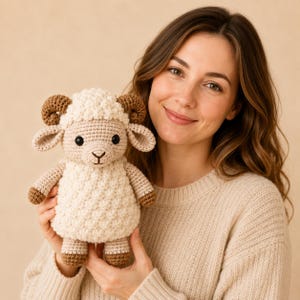 Puede incluir: Un animal de peluche tejido a ganchillo con forma de carnero, con cuerpo texturizado color crema, cara y extremidades de color tostado claro y cuernos curvos de color marrón oscuro. El carnero tiene dos ojos negros y nariz y boca bordadas.