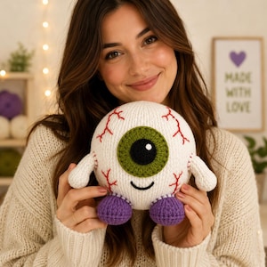May include: A round, white crocheted plush toy shaped like an eyeball with a large green iris, a black pupil, and a small black embroidered smile. The toy features red yarn veins stitched onto the white surface and two small purple feet.