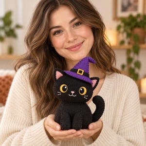 Puede incluir: Un peluche de gato hecho a mano en fieltro negro con grandes ojos amarillos, una pequeña nariz rosa y un sombrero de bruja morado con una hebilla amarilla. El gato tiene orejas rosas, bigotes blancos bordados y una cola curva de fieltro.
