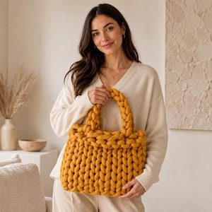 Può includere: Borsa tote lavorata a maglia grossa giallo senape con manici intrecciati. La borsa è realizzata in filato spesso e morbido e ha una forma rettangolare. La borsa è tenuta da una persona che indossa un maglione color crema.