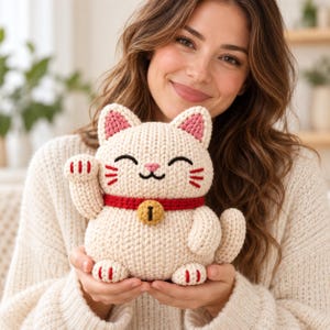 May include: A cream-coloured crocheted lucky cat figurine featuring a red collar with a gold-coloured bell, pink inner ears, and embroidered black facial features including eyes, a mouth, and whiskers, with one paw raised in a beckoning gesture.