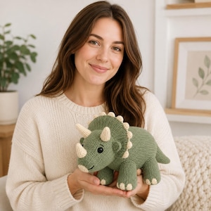 Puede incluir: Un juguete de triceratops tejido en color verde salvia con cuernos, garras y un borde de volante en color crema. El dinosaurio presenta un cuerpo redondeado, cuatro patas robustas, una cola corta y ojos negros, con una textura de punto visible.