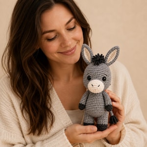 May include: A small, handmade crochet donkey toy featuring a grey body, a white muzzle, dark grey hooves, and a tuft of black hair on its head. The donkey has long, upright ears with light beige inner details and two small black safety eyes.