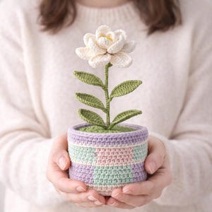 Könnte beinhalten: Eine gehäkelte Blume in einem gestreiften Topf. Die Blume ist weiß mit gelber Mitte, grünen Blättern und einem grünen Stiel. Der Topf ist mit Lavendel, Rosa und Mintgrün gestreift. Die handgefertigte Blume wird mit beiden Händen gehalten.