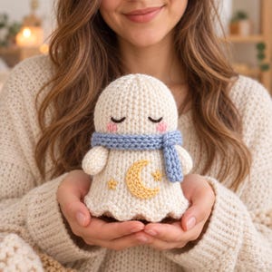 May include: A cream-coloured crocheted ghost plushie featuring a light blue scarf, embroidered closed eyes, pink cheeks, and a yellow crescent moon with two small stars on the front. The soft toy has a rounded head and a flared base design.