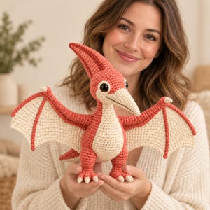 Puede incluir: Un juguete de pterodáctilo tejido a ganchillo con cuerpo y cabeza de color coral, además de vientre y membranas alares en color crema. El dinosaurio presenta una cresta prominente, grandes ojos oscuros y un pico puntiagudo de hilo.