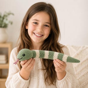 Peut inclure: Un serpent en tricot avec des rayures alternées vert sauge et crème. Le serpent possède une tête arrondie avec des yeux en boutons noirs, un petit sourire brodé et une langue fourchue en fil rose dépassant de sa bouche.