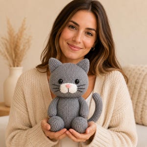 May include: A grey knitted cat plush toy featuring a white muzzle, a small pink nose, black embroidered whiskers, and two round black eyes. The stuffed animal has a rounded body, two front paws, two back paws, and a curved tail extending to the side.