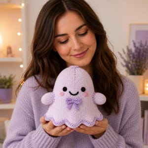 Pode incluir: Uma peluche de fantasma tricotada em violeta claro com um rosto sorridente, olhos bordados a preto, uma pequena boca preta e duas bochechas circulares cor-de-rosa. O fantasma tem um pequeno laço violeta ao centro e uma orla recortada.