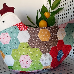 May include: A handcrafted chicken-shaped basket made of colorful fabric hexagons. The basket features a white head, red comb, and a black eye. The basket is filled with green leaves and two small oranges.