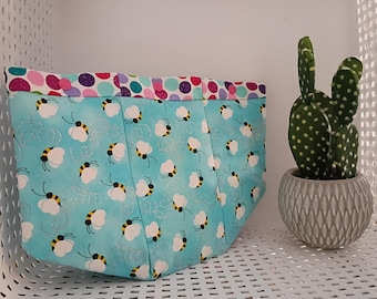 May include: A turquoise fabric tote bag with a pattern of white bees with black and yellow details. The top edge has a colorful polka dot pattern. A small cactus in a gray pot sits nearby.