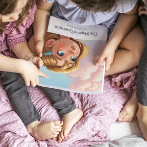 May include: A children's book titled "The Magic of Questions" is held by three children. The book cover features a cartoon illustration of a girl with blonde hair and blue eyes. The children are sitting on a pink floral patterned blanket.