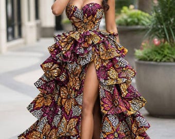 May include: A strapless floor-length gown featuring a vibrant floral pattern in shades of purple, orange, and white. The dress has a fitted bodice and a voluminous skirt composed of multiple ruffled layers with a high front slit revealing the leg.